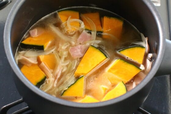 炒めたベーコンと玉ねぎが入った鍋に、かぼちゃ、水、顆粒コンソメを加える